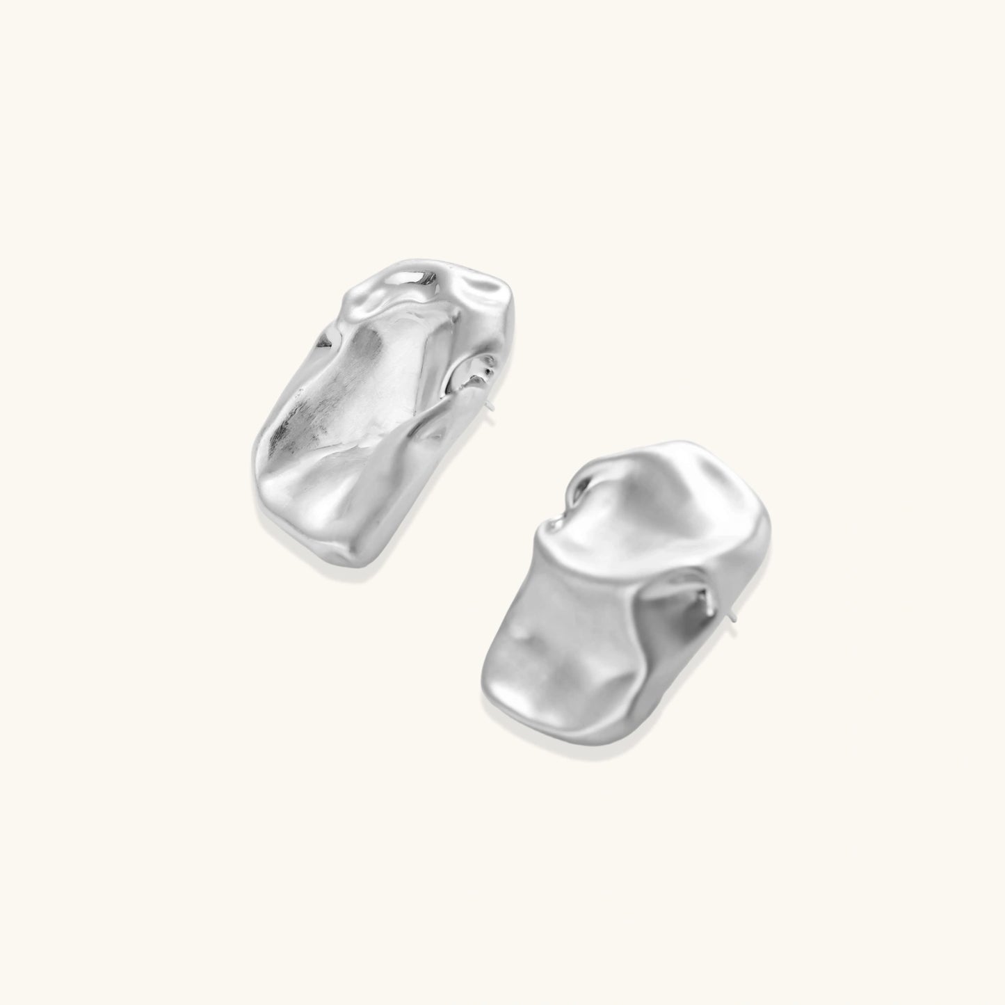 Pair of sculptural silver ear cuffs with organic fluid shapes and polished finish against cream background.