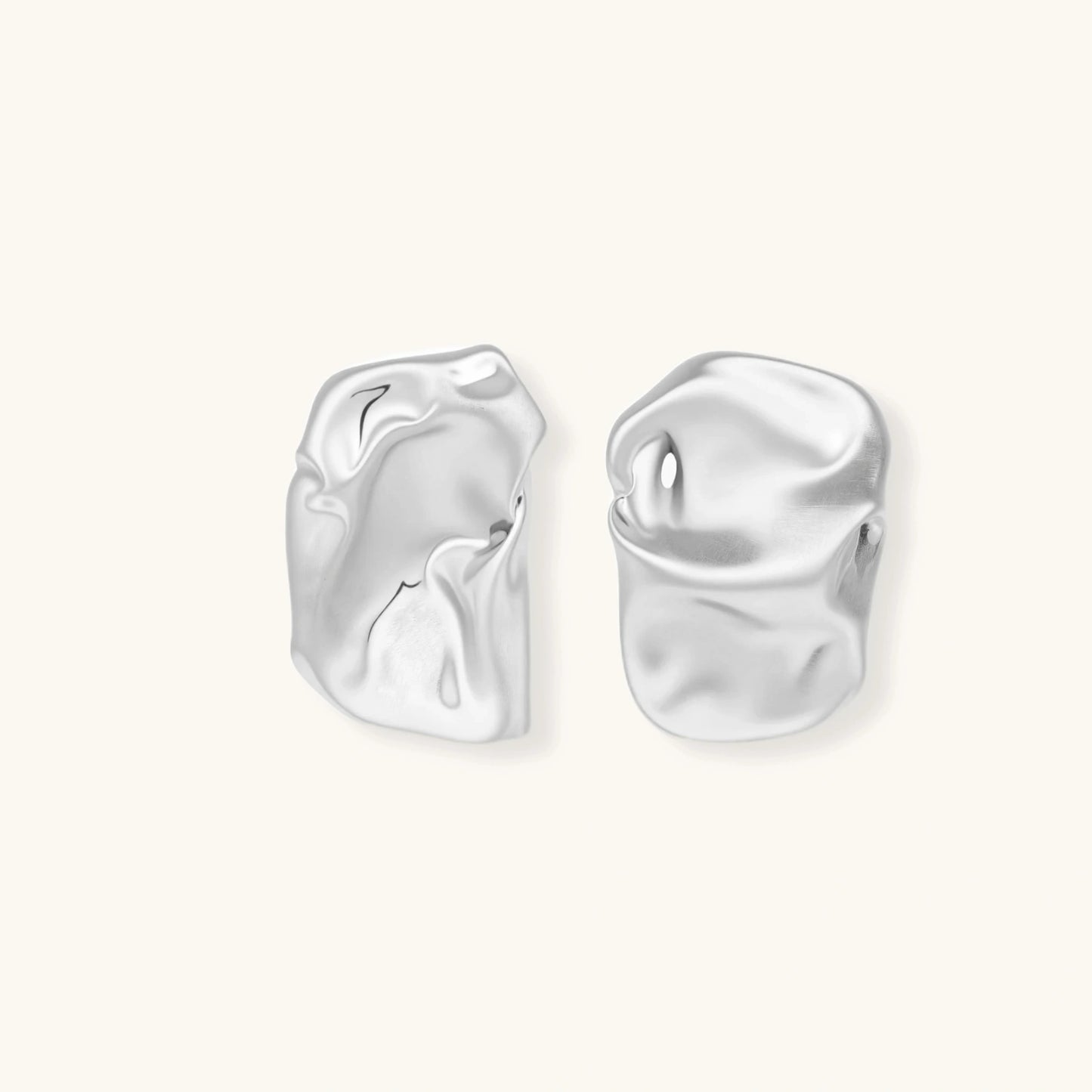 Pair of sculptural silver ear cuffs with organic fluid shapes and polished finish against cream background.